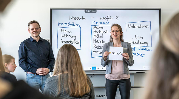 Seminarszene: Zwei Vortragende vor einer Tafel erklären, wofür Kredite genutzt werden.